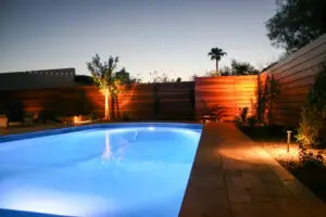 Backyard pool area at dusk with blue pool lights illuminating the water and warm landscape lighting highlighting a modern wooden fence and a small tree.