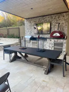 Stylish outdoor kitchen area with a sleek dining table, built-in grill, pizza oven, and a wall-mounted TV, perfect for entertaining guests.