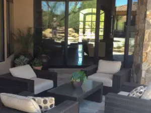 Cozy patio area with cushioned wicker chairs, a glass-top coffee table, potted plants, and large glass doors reflecting the outdoor landscape.