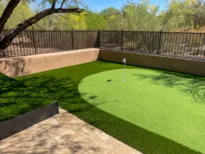 Outdoor putting green with lush artificial grass, bordered by a wrought iron fence and natural desert landscape.