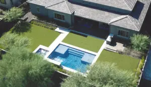 Aerial view of a backyard with a modern rectangular swimming pool and a spa, surrounded by green artificial turf and a paved patio area adjacent to the house.