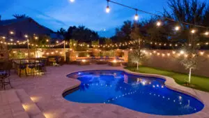 Night view of a backyard with a swimming pool, string lights, outdoor bar with high chairs, and a cozy seating area with fire pits, creating a warm and inviting atmosphere.