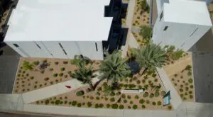 Aerial view of an apartment complex with flat roof, surrounded by landscaped pathways and desert vegetation, including palm trees and various shrubs.