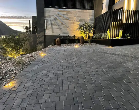 Close-up of a modern driveway with interlocking pavers and landscape lighting leading to a contemporary hillside home with desert landscaping.