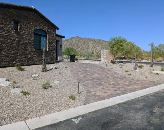 Paved driveway with a rustic brick design leading to a stone house in a desert landscape.