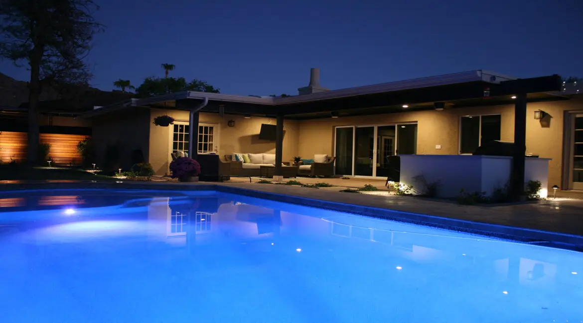 Night view of a modern poolside lounge with illuminated pool, cozy seating area, outdoor kitchen, and ambient lighting.