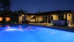 Night view of a modern poolside lounge with illuminated pool, cozy seating area, outdoor kitchen, and ambient lighting.