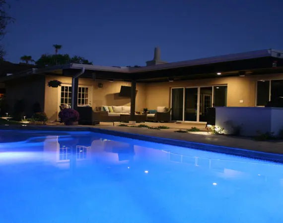 Night view of a modern poolside lounge with illuminated pool, cozy seating area, outdoor kitchen, and ambient lighting.