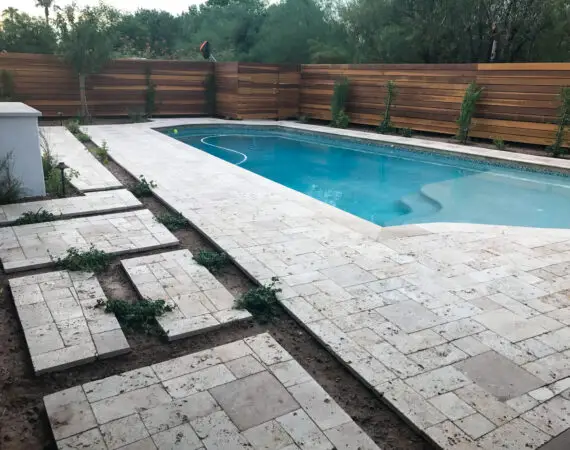 Elegant backyard with a modern pool, travertine pavers, and wooden privacy fence, featuring lush greenery and landscaped planters.