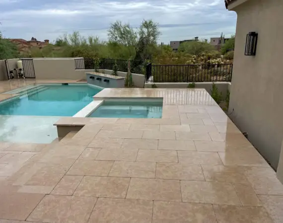 Modern pool and spa with sleek, light-colored stone decking, surrounded by black iron fencing and natural desert landscaping.