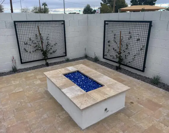 Rectangular fire pit with blue glass stones in a light stone patio area, surrounded by a white wall and trellis with climbing plants.