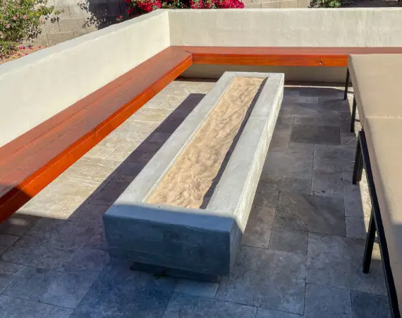 Modern concrete fire pit with a linear design, surrounded by built-in wooden benches and a stone patio, set against a brick wall with blooming bougainvillea.