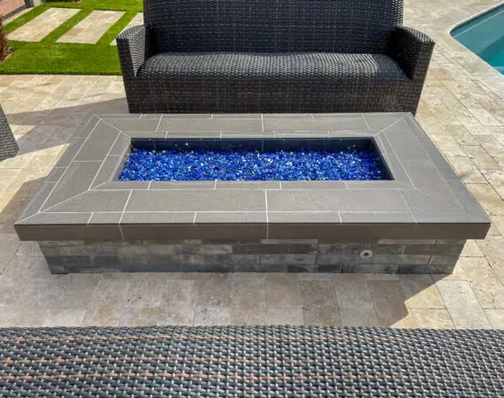 Modern rectangular fire pit with blue glass stones, surrounded by wicker seating on a stone patio next to a pool.