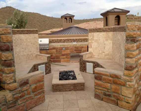 Outdoor kitchen with built-in grill, stone fire pit, and seating area made from natural stone and tile, set against a backdrop of desert hills.