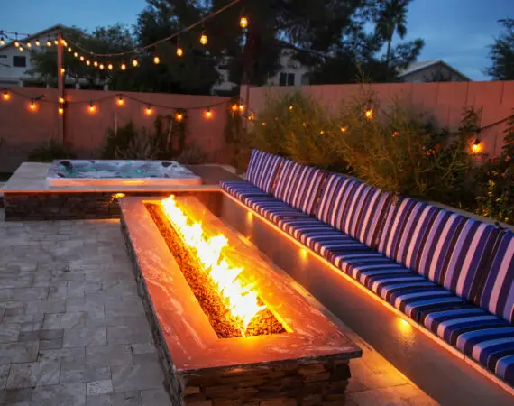 Modern backyard fire feature with a long rectangular fire pit, stone base, and blue striped bench seating, illuminated by string lights at dusk.