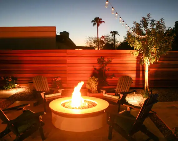 Cozy backyard fire pit area with Adirondack chairs, surrounded by string lights and a modern wooden fence, creating a warm evening ambiance.