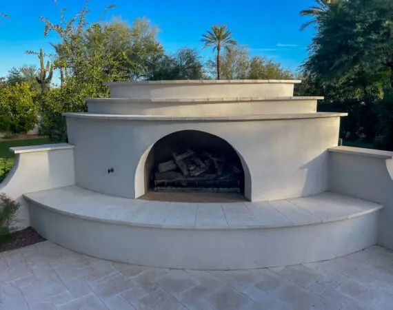 Large circular outdoor fireplace with a tiered design and open firebox, set on a spacious stone patio, surrounded by lush greenery and desert plants.