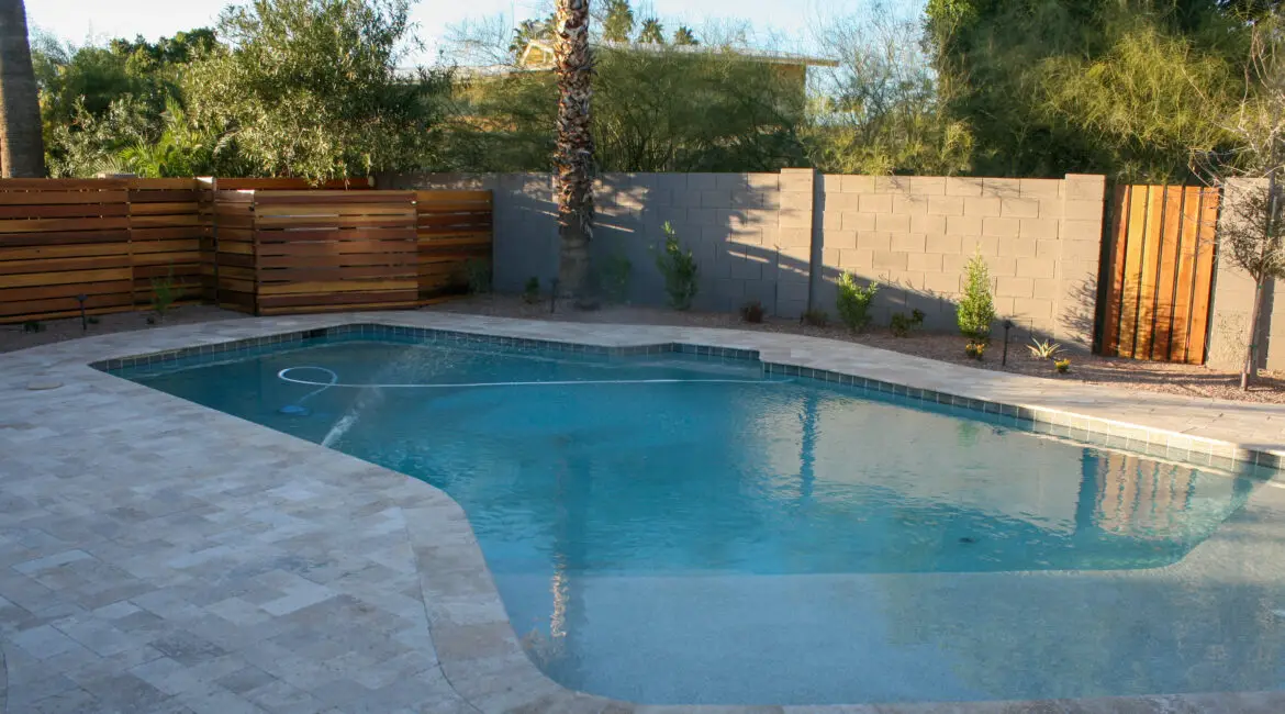 Backyard swimming pool with a modern travertine deck, wooden privacy fence, and lush greenery providing a serene and inviting atmosphere.