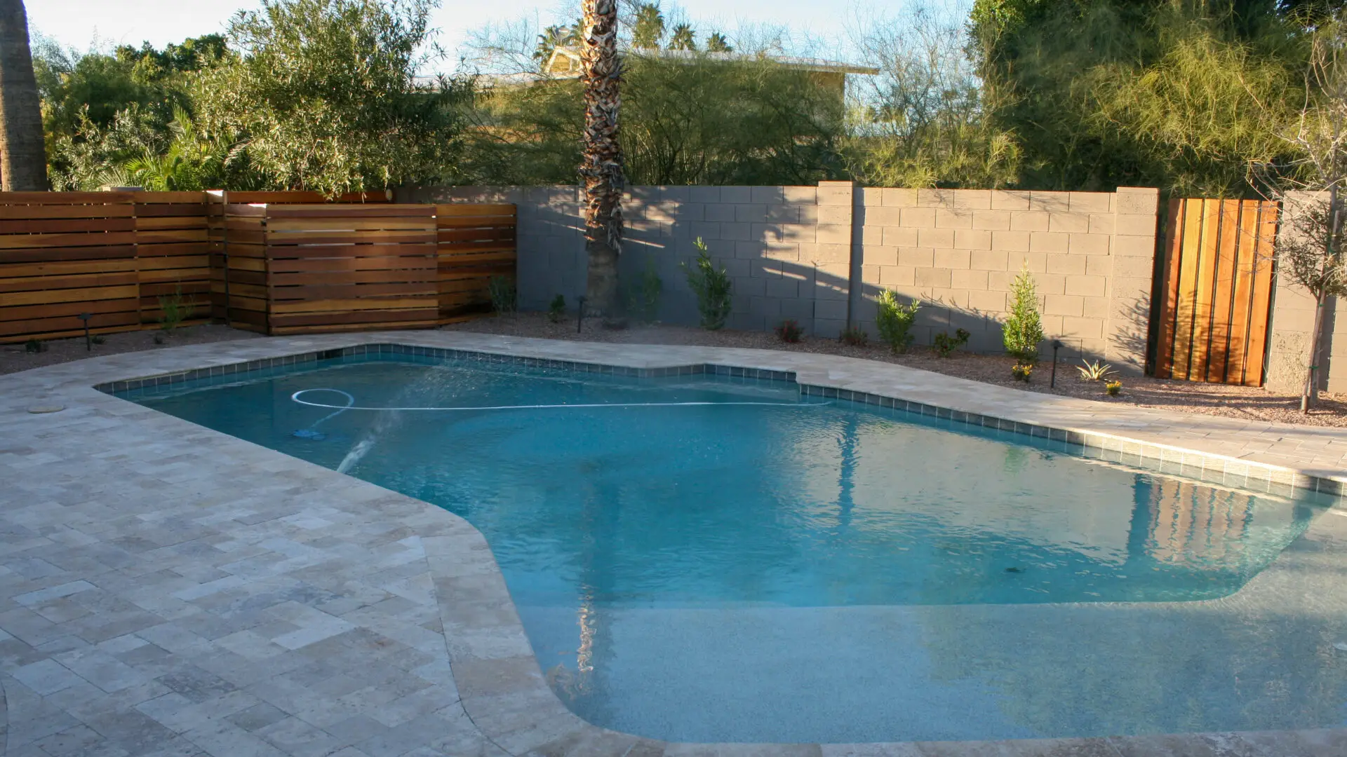 Backyard swimming pool with a modern travertine deck, wooden privacy fence, and lush greenery providing a serene and inviting atmosphere.