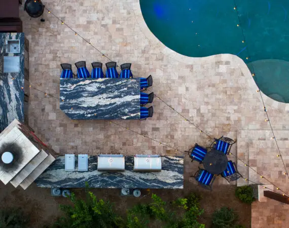 Aerial view of an outdoor kitchen and dining area with marble countertops, string lights, and a swimming pool.