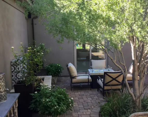 Cozy courtyard with cushioned seating, potted plants, and brick paving, shaded by a tree.