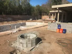 Construction site of a backyard renovation project featuring an unfinished fire pit and swimming pool, with materials and tools scattered around.