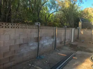 Backyard construction site with a partially completed cinder block wall, metal poles, and construction materials.