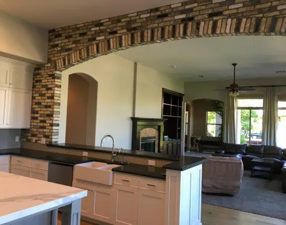 Modern kitchen with white cabinets, farmhouse sink, marble island, and brick archway leading to a cozy living area.