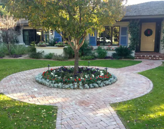 Front yard of a house with a brick walkway, a tree surrounded by colorful flowers, and well-maintained grass, with the house entrance decorated with a wreath.