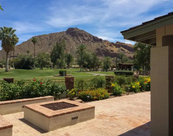 View of a landscaped yard with a brick patio, planters, and vibrant flowers, with a backdrop of green fields and a mountain.