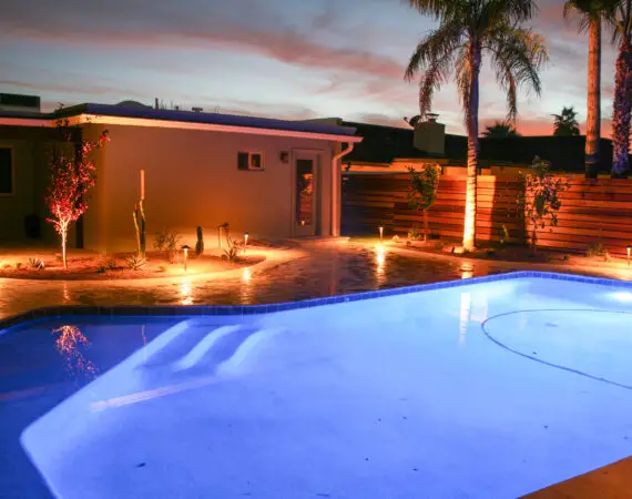 Backyard pool area illuminated with vibrant lighting, showcasing a landscaped garden and palm trees against a twilight sky.