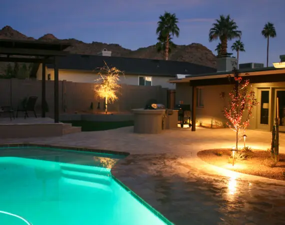 Backyard with illuminated pool and garden, featuring strategically placed landscape lights that highlight a tree and patio area at dusk.