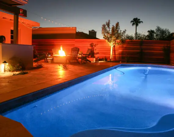 Backyard pool area with blue pool lights and a lit fire pit surrounded by Adirondack chairs at dusk, featuring landscape lighting and a modern wooden fence.