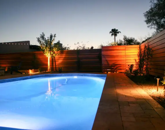 Backyard pool area at dusk with blue pool lights illuminating the water and warm landscape lighting highlighting a modern wooden fence and a small tree.