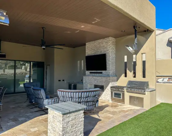 Modern outdoor patio featuring a dining area with a long table and chairs, a built-in grill, a pizza oven, and a mounted TV on a stone wall.