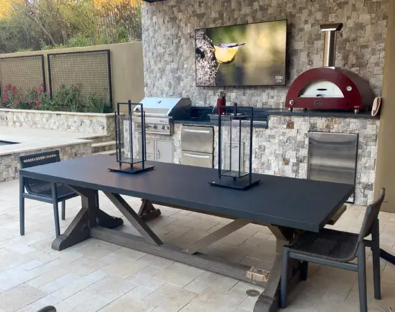 Outdoor patio area featuring a sleek dining table, modern chairs, a built-in grill, and a TV mounted on a stone wall with decorative lanterns.