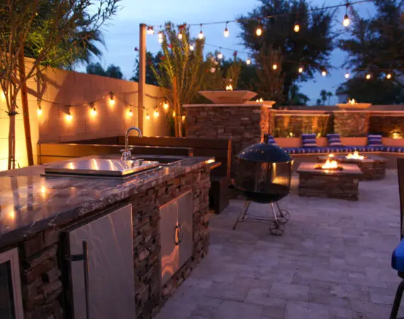 Elegant outdoor kitchen with stone counters, a built-in sink, and grill, surrounded by string lights and a cozy fire pit lounge area.