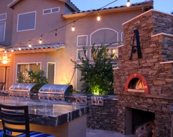 Elegant outdoor kitchen setup with a large marble counter, built-in grills, a stone pizza oven, and string lights for ambient lighting.