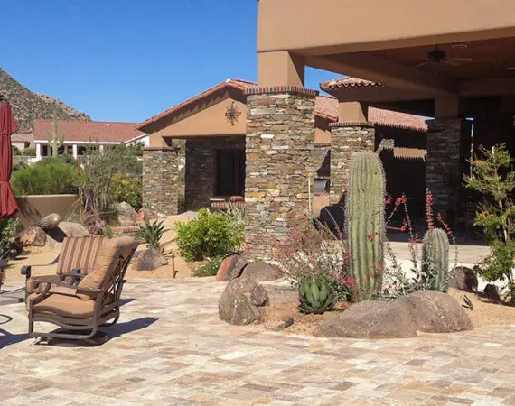 Desert-themed patio with stone pillars, a seating area with cushioned chairs, cacti, and a paved surface against a mountain backdrop.