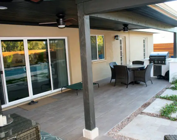 Modern outdoor patio featuring sliding glass doors, ceiling fans, a dining set, a grill, and a paved walkway leading to a pool area.