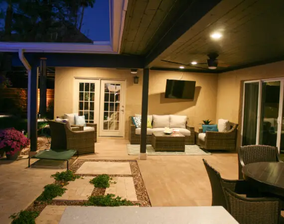 Evening view of an outdoor patio featuring wicker furniture, a wall-mounted TV, decorative plants, and a paved walkway, creating a cozy ambiance.