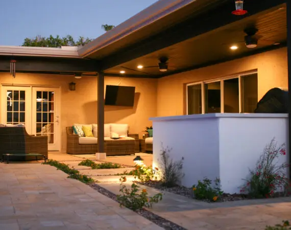 Cozy outdoor patio with wicker furniture, a wall-mounted TV, barbecue area, and decorative plants, providing a perfect space for evening gatherings.