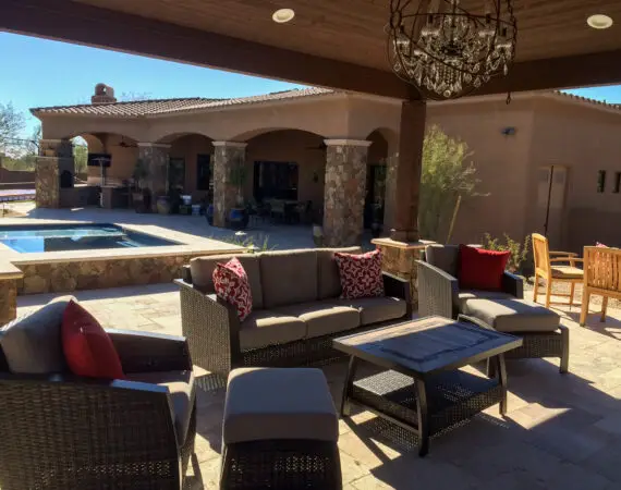 Luxurious outdoor patio with comfortable wicker furniture, decorative cushions, a chandelier, and a pool area with stone pillars and arches in the background.