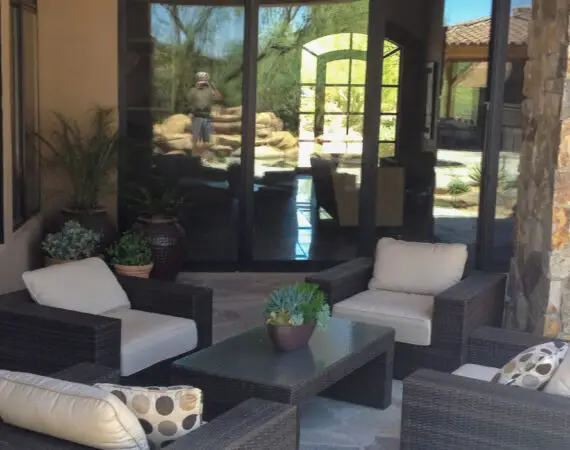 Cozy patio area with cushioned wicker chairs, a glass-top coffee table, potted plants, and large glass doors reflecting the outdoor landscape.