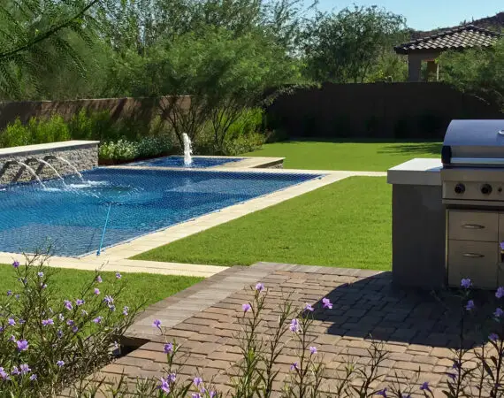 Beautiful backyard with a pool, water features, built-in grill, and lush green lawn with purple flowers.