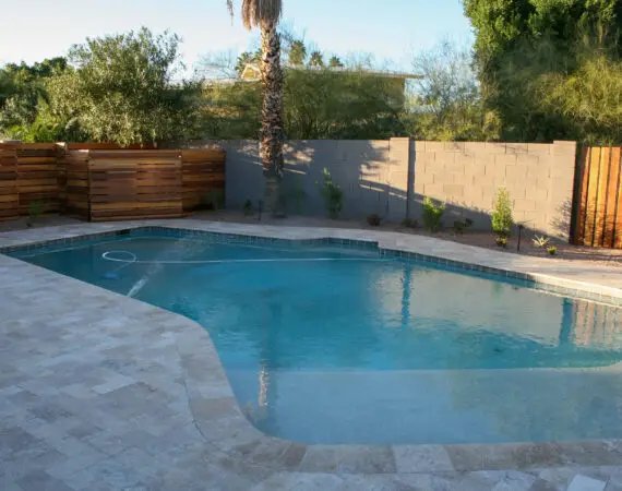 Backyard swimming pool with clean, clear water and a stone tile deck, surrounded by a privacy fence and lush greenery.