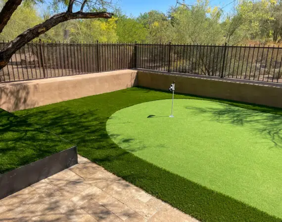 Outdoor putting green with lush artificial grass, bordered by a wrought iron fence and natural desert landscape.