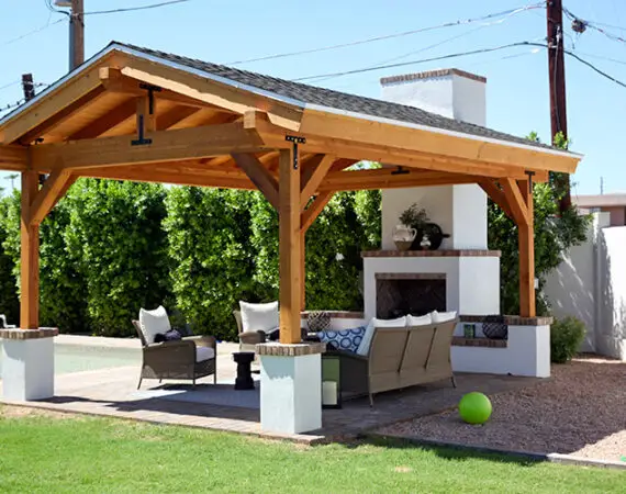 Cozy outdoor patio with a wooden pavilion, featuring a stone fireplace, comfortable seating, and a lush green lawn.