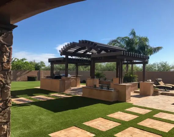 Outdoor kitchen with a large wooden pergola, stone countertops, and built-in grills, set on a lush lawn with stone pathways and lounge chairs.