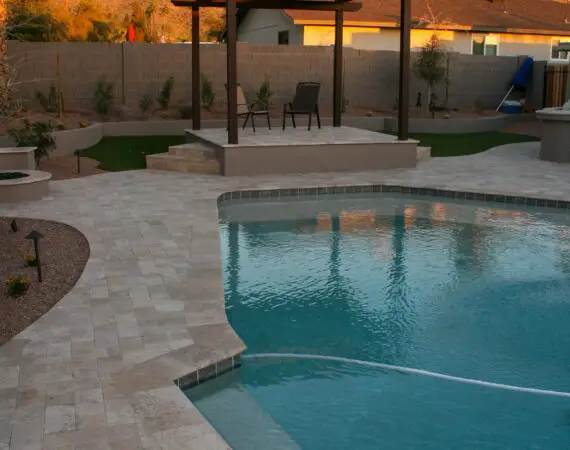 Backyard pool area with a wooden pergola and stone pathway, featuring desert landscaping and mountain views at sunset.